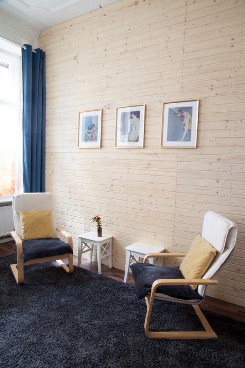 Zwei weiße Sessel mit Kissen stehen sich gegenüber vor einer hellen Holzwand. An der Wand hängen drei gerahmte Bilder. Zwischen den Sesseln stehen zwei kleine weiße Tische an der Wand.